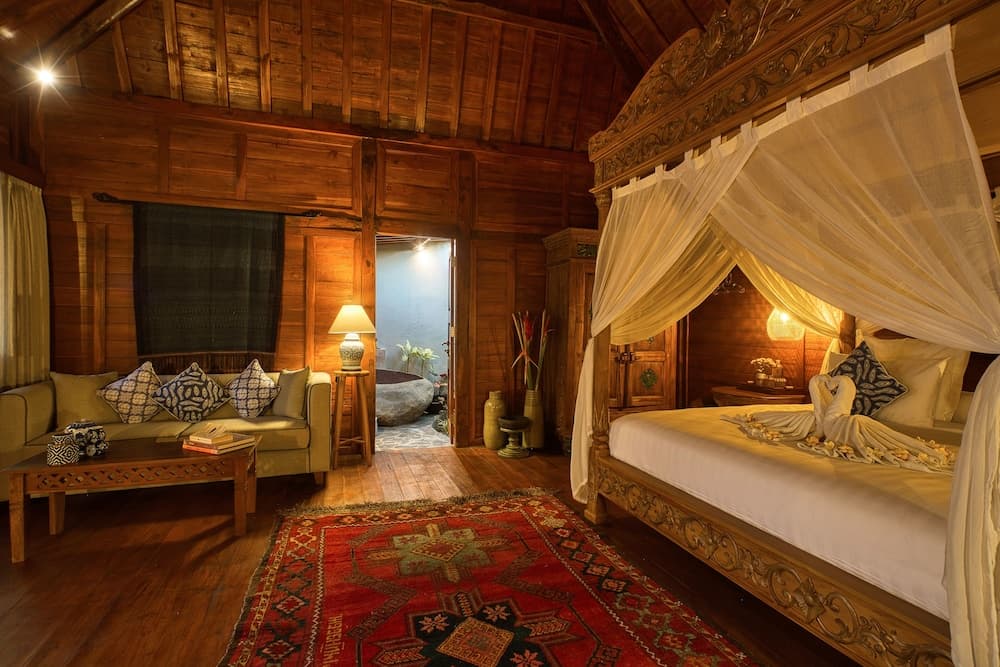 A cozy wooden cabin bedroom features a carved canopy bed draped in sheer curtains, a vibrant red rug, and warm lighting, creating an inviting retreat ideal for travelers seeking rustic luxury.