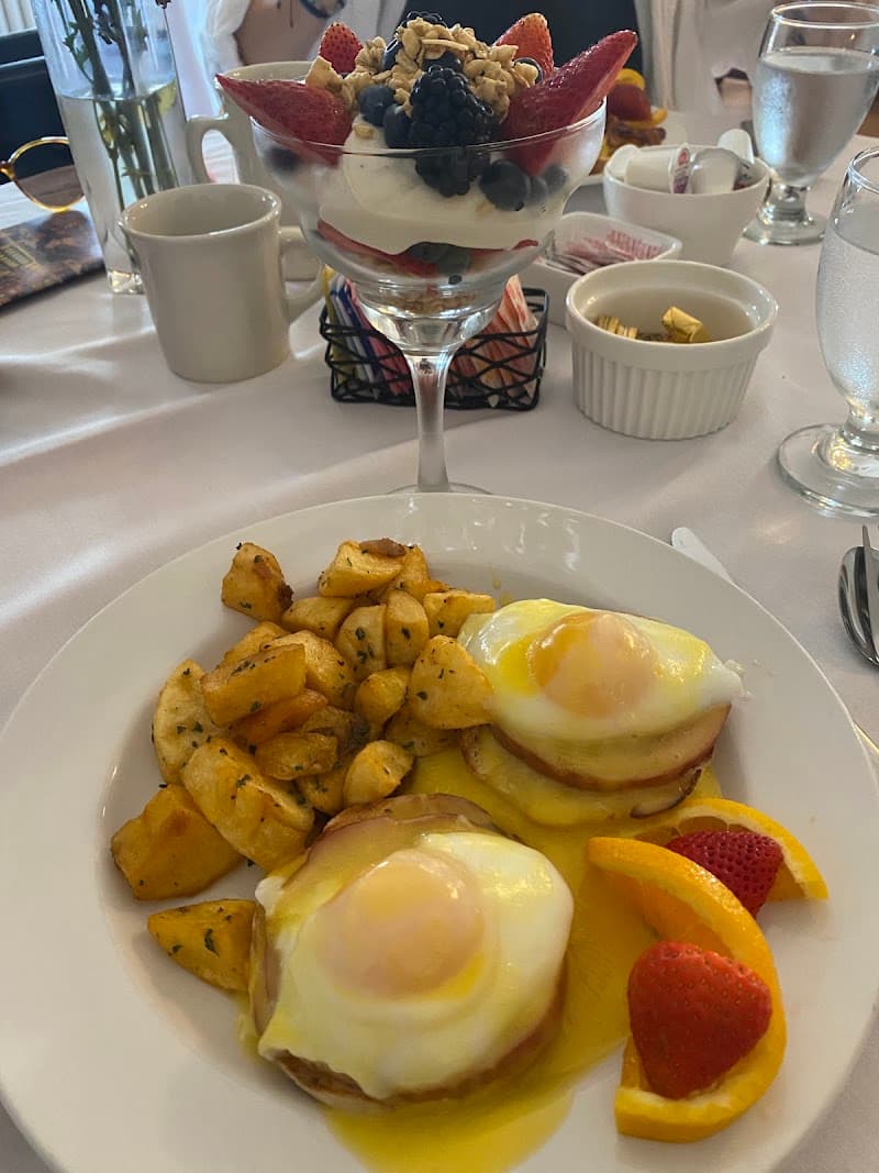 A cozy breakfast scene at the Castle Manor Inn in Gloucester, MA, features sunny-side-up eggs benedict, crispy potatoes, fresh fruit, and a vibrant parfait, creating an inviting and indulgent dining experience perfect for travelers seeking comfort food.