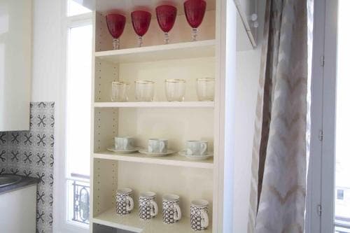 A cozy kitchen corner features a neatly organized shelf of colorful mugs and glasses, set against a backdrop of patterned curtains and a bright window, ideal for travelers seeking a charming and homey accommodation.