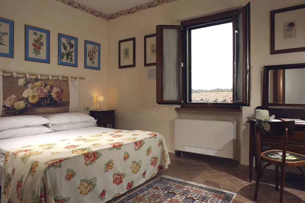 A cozy bedroom with floral bedding, framed botanical prints, and an open window offering a serene view, ideal for travelers seeking a tranquil rural retreat.