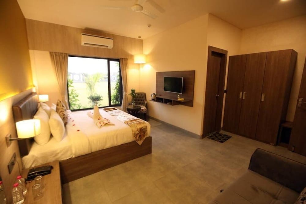 A cozy hotel room with warm lighting, a neatly made bed adorned with decorative towels, a flat-screen TV, and a view of greenery through large windows, ideal for travelers seeking comfort and relaxation.