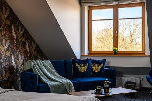 A cozy attic room with a plush blue sofa adorned with golden-embroidered pillows, a patterned wallpaper wall, and a bright window showcasing leafless trees, ideal for a serene getaway or a romantic retreat.
