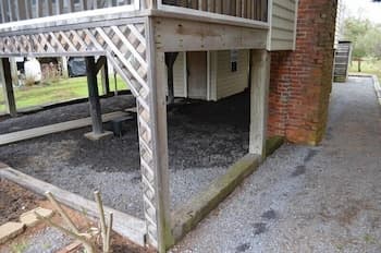 A rustic wooden deck supported by lattice pillars casts shade over a gravel area beside a brick building, evoking a serene suburban atmosphere ideal for exploring quaint neighborhoods.