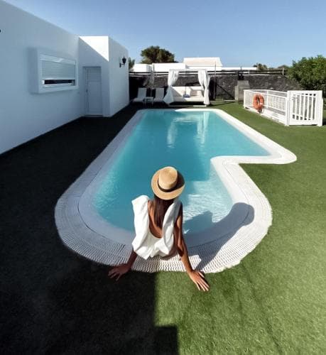 A serene vacation scene features a woman relaxing by a modern poolside on artificial turf, surrounded by minimalist architecture and outdoor furniture, ideal for luxury travel content.