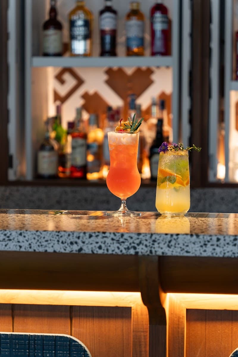 A vibrant bar scene at SAii Laguna Phuket features two beautifully garnished cocktails on a sleek countertop, set against a backdrop of illuminated liquor bottles, creating an inviting atmosphere perfect for travelers seeking a luxurious tropical retreat.