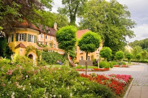 A serene European village scene features charming yellow buildings nestled among lush greenery and vibrant flower beds, ideal for travelers seeking picturesque countryside tranquility.
