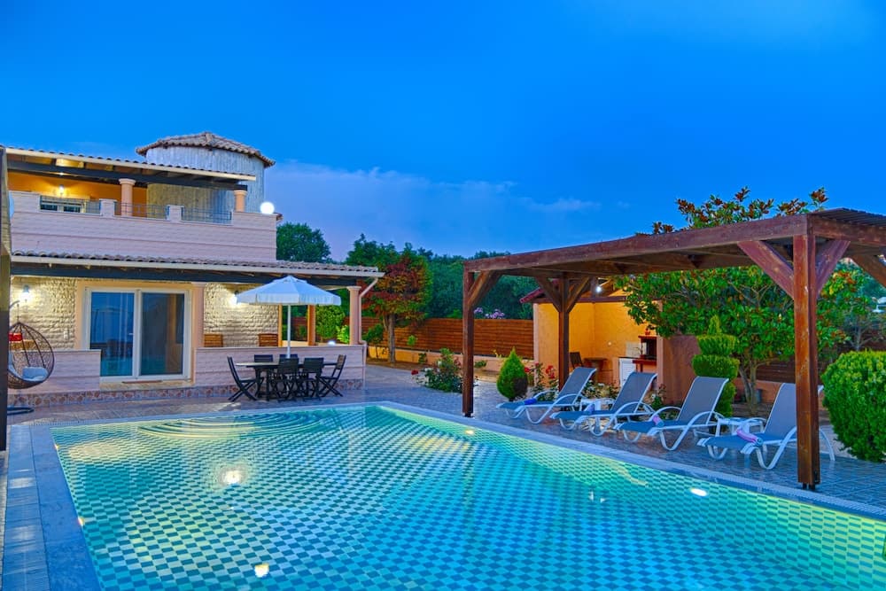 A serene evening at a luxurious villa features a glowing pool, cozy lounge chairs under a wooden pergola, and a dining area with an umbrella, ideal for a tranquil vacation retreat.