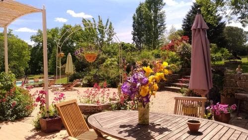 A serene garden patio features vibrant flowers, wooden furniture, and a closed umbrella under a sunny sky, ideal for relaxing getaways or romantic retreats.