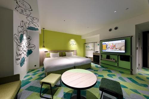 A vibrant hotel room features playful green and white decor, a cozy bed, interactive wall art, and a modern entertainment setup, ideal for travelers seeking a fun and relaxing stay.