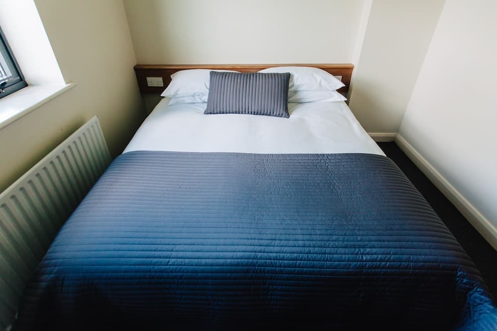 A cozy bedroom with a neatly made bed featuring a blue quilted cover and white pillows, set against light walls with a window and radiator, ideal for travelers seeking a comfortable and minimalist accommodation.