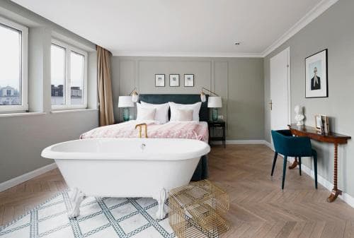 A cozy hotel room features a stylish clawfoot bathtub in the center, soft gray walls, a neatly made bed by large windows, and a chic desk area, ideal for travelers seeking comfort and elegance.