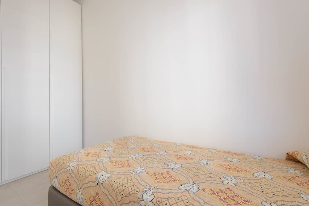 A cozy bedroom corner features a neatly made bed with a floral-patterned quilt, white walls, and a minimalist wardrobe, ideal for travelers seeking simple and clean accommodations.
