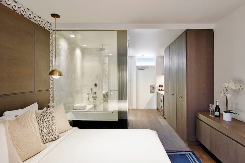 A cozy hotel suite at the Kaskades Hotel South Beach features a modern bedroom with plush bedding, an open bathroom with a luxurious bathtub, and sleek wooden furnishings, ideal for travelers seeking comfort and style in Miami Beach.