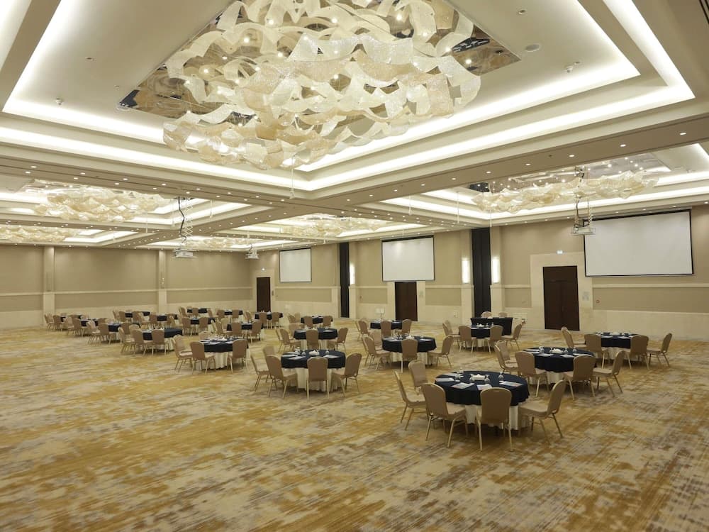 A spacious conference hall with elegant chandeliers, round tables set for dining, and large screens, ideal for corporate events or weddings.