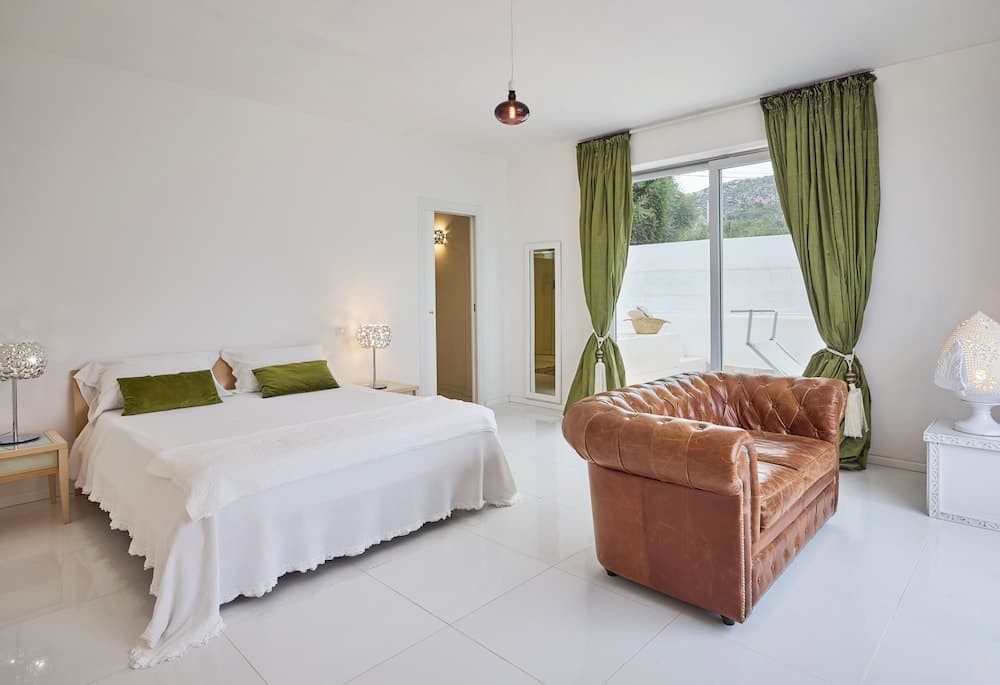 A serene bedroom with white walls, a cozy bed adorned with green pillows, a leather armchair, and large windows framed by lush green curtains offers a tranquil retreat ideal for vacation rentals.