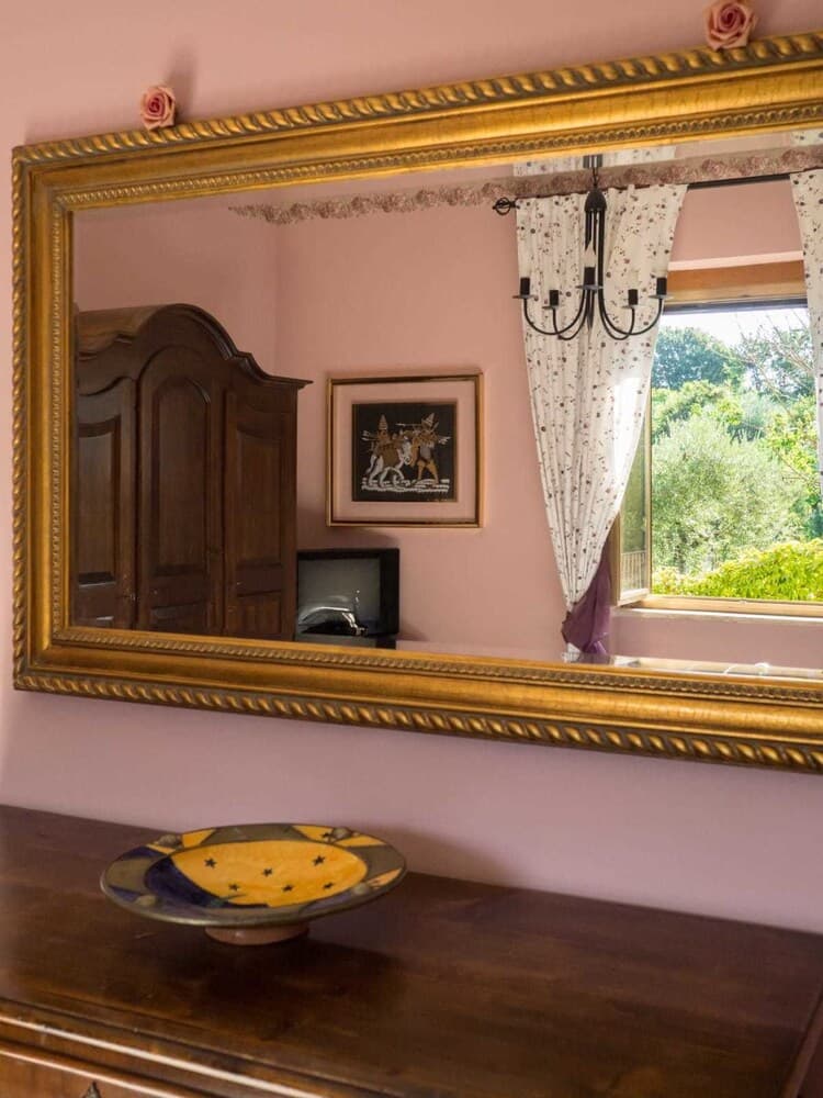A cozy bedroom with soft pink walls, a large ornate mirror reflecting a wooden wardrobe and framed artwork, a decorative plate on a dresser, and a window with curtains framing lush greenery outside, ideal for travelers seeking a charming and serene retreat.