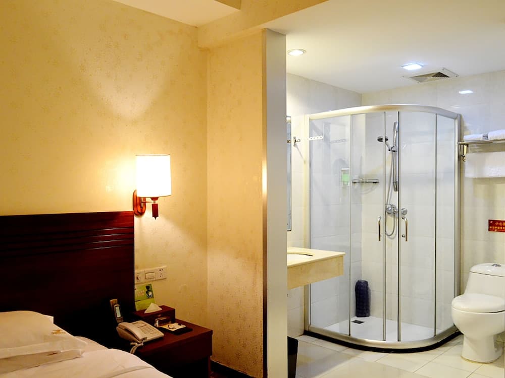 A cozy hotel room features a neatly made bed beside a warmly lit bedside table, adjacent to a modern bathroom with a glass-enclosed shower, ideal for travelers seeking comfort and convenience.