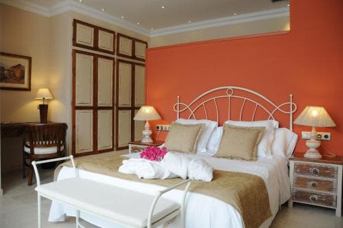 A cozy hotel room features a neatly made bed adorned with pillows and flowers, set against an orange accent wall, complemented by wooden furnishings and bedside lamps, ideal for travelers seeking comfort and relaxation.