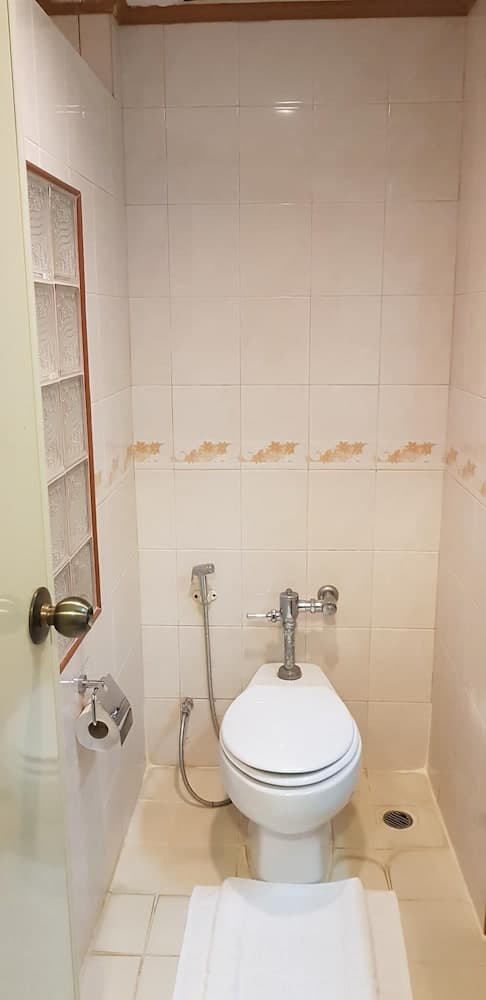 A compact bathroom with beige tiled walls, a white toilet, bidet hose, and frosted glass block window offers a functional and clean space ideal for travelers seeking basic amenities in budget accommodations.