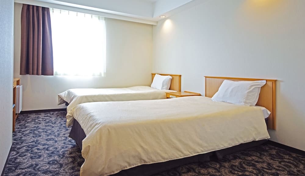 A cozy hotel room features two neatly made beds with wooden headboards, soft lighting, and a large window with curtains, ideal for travelers seeking comfort and simplicity.