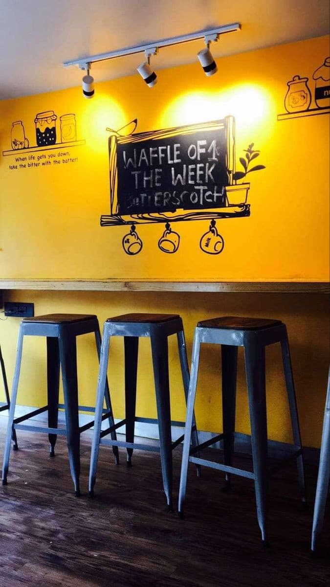 A cozy café corner with vibrant yellow walls adorned with playful chalkboard art and jars, featuring metal stools against a wooden counter, perfect for travelers seeking a charming spot to enjoy a waffle of the week in Sadar Bazaar, Meerut.