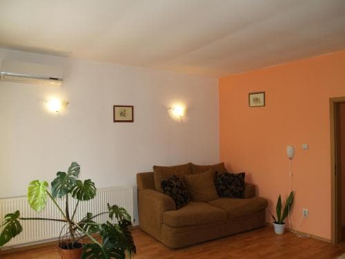 A cozy living room with warm orange walls, a comfortable sofa adorned with patterned cushions, lush green plants, and soft lighting creates an inviting atmosphere ideal for travelers seeking a relaxing accommodation.