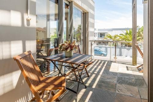 A sunlit balcony with wooden furniture and potted plants overlooks a serene coastal area, ideal for travelers seeking relaxation in a modern vacation rental.