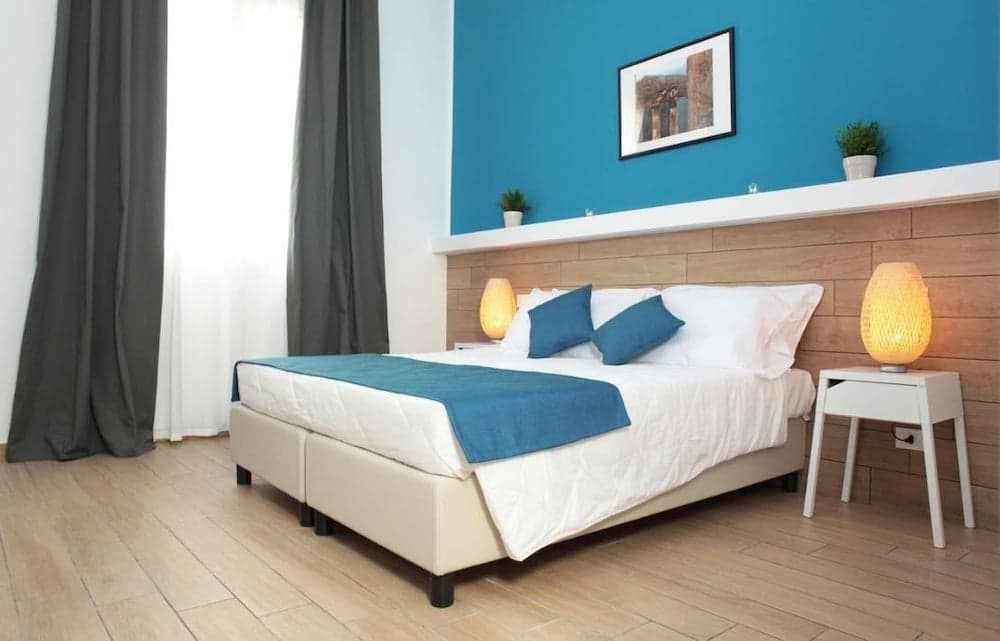 A cozy hotel bedroom features a neatly made bed with blue accents against a vibrant blue wall, wooden flooring, and soft lighting, ideal for travelers seeking comfort and relaxation.