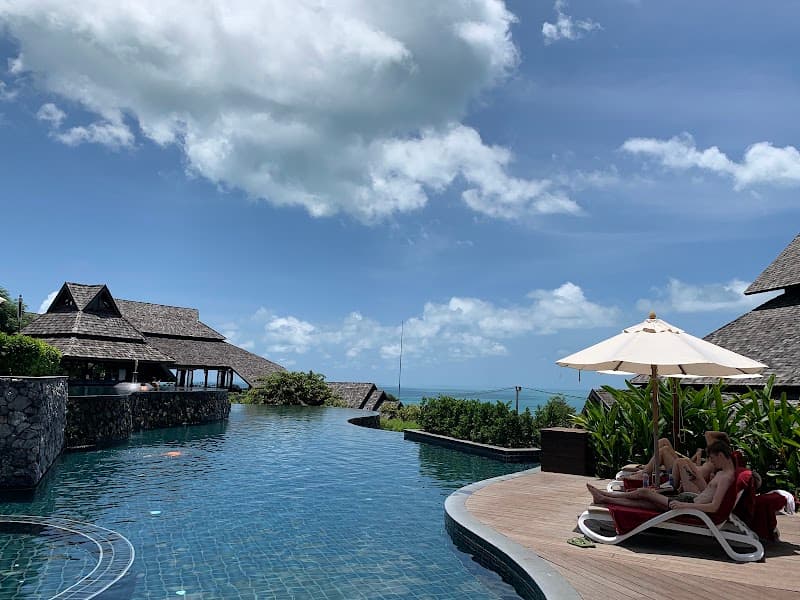 A serene tropical resort scene features a tranquil infinity pool overlooking lush greenery and the ocean under a partly cloudy sky, with guests relaxing on lounge chairs under umbrellas, ideal for luxury beachfront vacations in Koh Samui, Thailand.