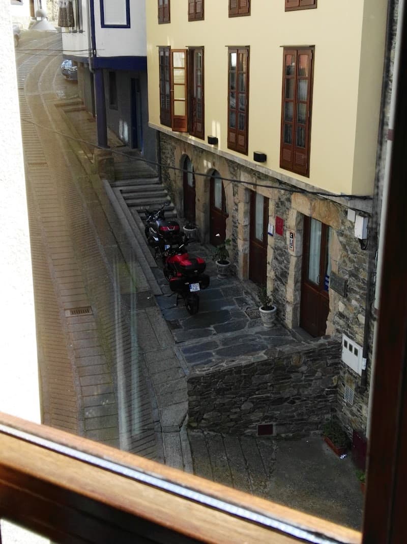 A cozy, sunlit view of a quaint Spanish street in Asturias features stone buildings, wooden shutters, parked motorcycles, and potted plants, ideal for travelers seeking a charming European getaway experience.