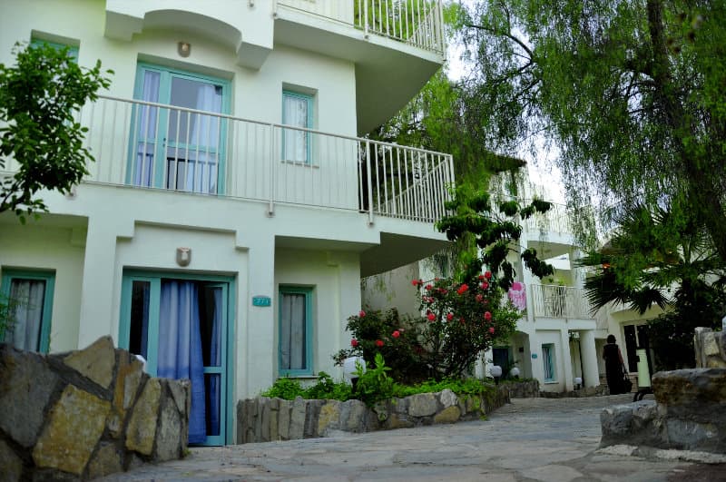 A serene stone-paved pathway winds through lush greenery beside a charming white building with turquoise accents, ideal for travelers seeking a tranquil retreat in a picturesque setting.