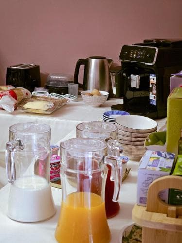 A cozy breakfast buffet setup featuring pitchers of milk and orange juice, stacked plates, a coffee machine, and assorted breakfast items on a tablecloth-covered table, ideal for travelers seeking a warm morning start in a hotel or guesthouse.