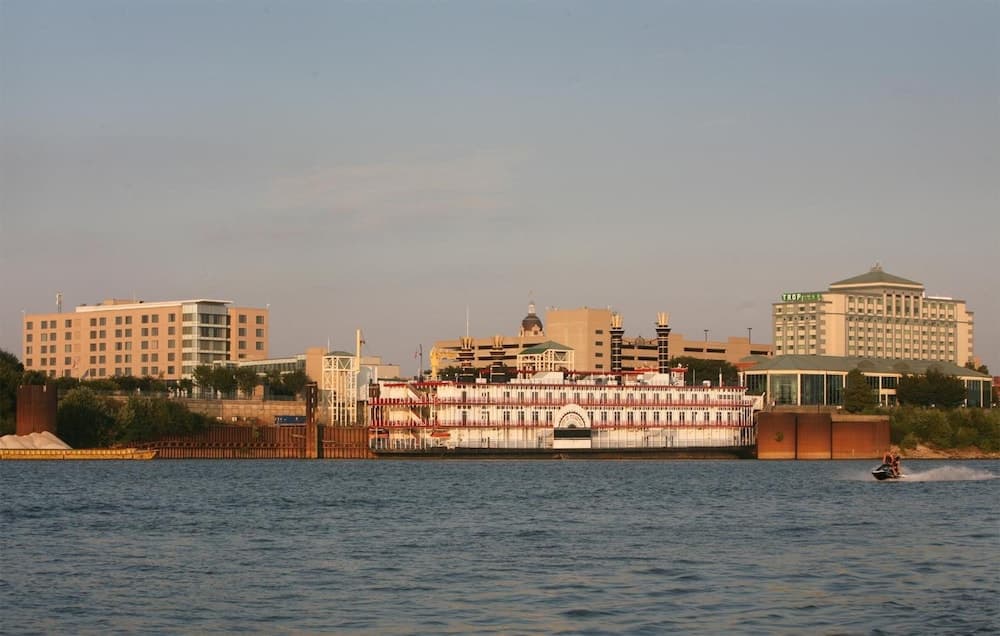 A serene waterfront cityscape features a paddlewheel riverboat docked near modern buildings, with a jet ski adding motion to the calm waters, ideal for travelers seeking urban riverfront adventures.