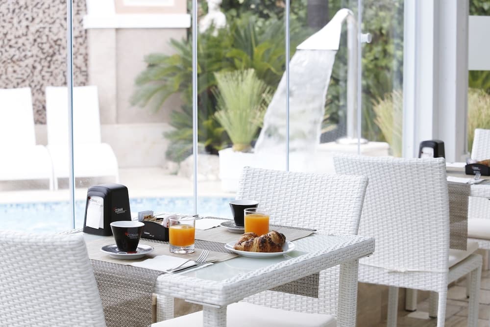 A serene hotel patio features white wicker furniture set with breakfast items near a glass-enclosed pool area, ideal for travelers seeking a relaxing morning ambiance.