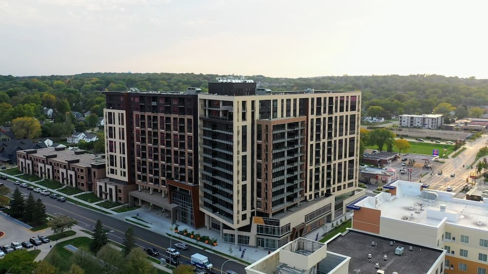 A modern high-rise apartment building stands prominently amidst lush greenery and suburban homes, offering an ideal urban retreat with scenic views, perfect for city dwellers seeking a blend of nature and convenience.