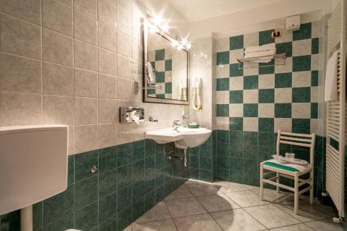 A cozy hotel bathroom features green and white checkered tiles, a sink with a mirror, a towel rack, and a wooden chair, creating a clean and inviting atmosphere ideal for travelers seeking comfort.