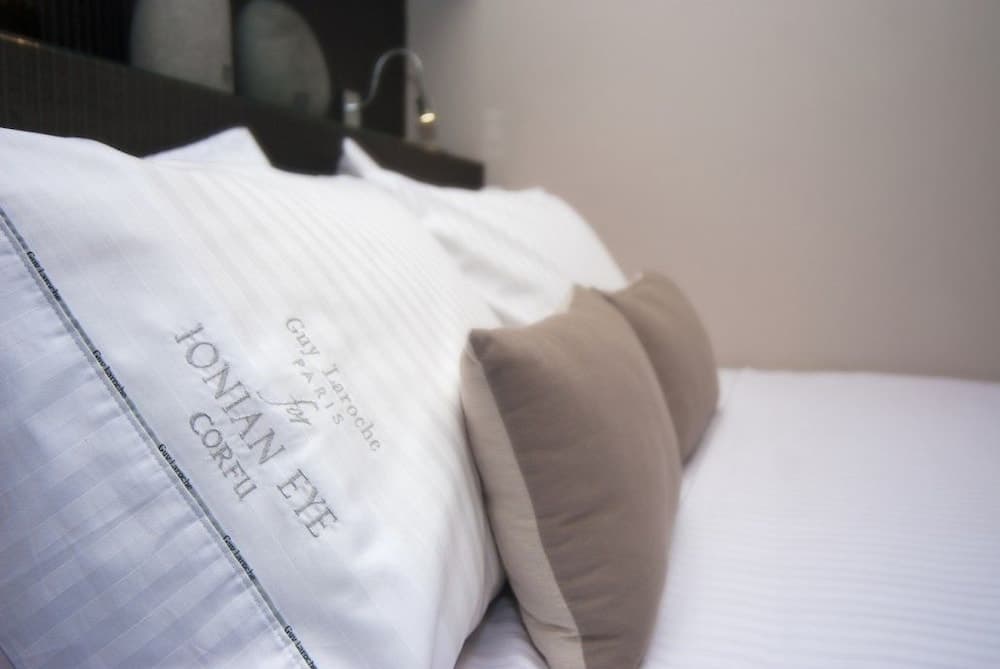 A cozy hotel room bed adorned with elegant "Ionian Eye" pillows invites travelers to unwind in serene comfort, perfect for a relaxing getaway.