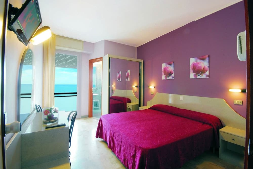 A cozy hotel room with vibrant purple walls, red bedding, and ocean views through a balcony window offers a serene retreat for travelers seeking relaxation by the sea.