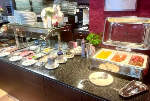A well-lit hotel buffet station showcases an array of fresh breakfast items like fruits, cheeses, and spreads, inviting travelers to start their day with a hearty meal in a cozy dining area.