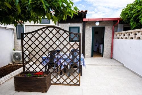 A cozy outdoor patio features a lattice screen, potted flowers, and a table set for dining under a leafy tree, creating an inviting atmosphere ideal for travelers seeking a tranquil accommodation experience.