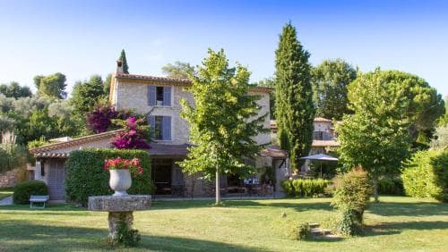 A serene countryside villa surrounded by lush greenery and vibrant flowers creates an inviting atmosphere, perfect for a tranquil getaway, featuring a charming fountain and shaded seating areas ideal for relaxation.
