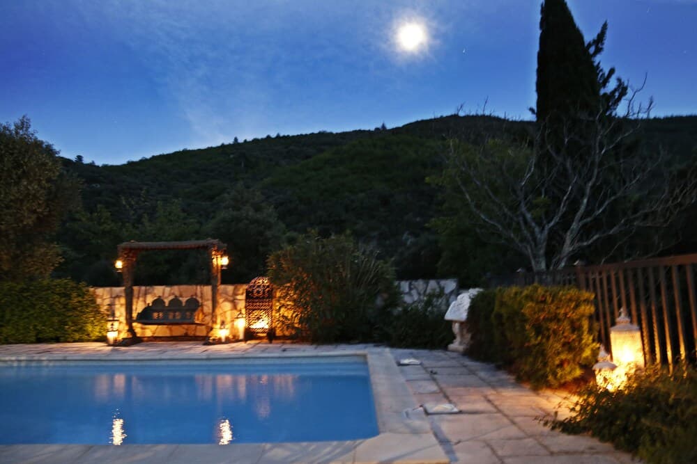 A serene evening at a luxurious villa features a tranquil pool illuminated by warm lanterns under a full moon, surrounded by lush greenery and hills, ideal for romantic getaways or peaceful retreats.