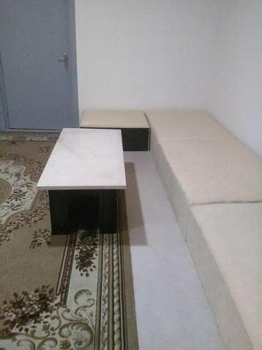 A cozy indoor seating area features a sleek white table on a patterned rug, complemented by beige cushioned benches against a minimalist wall, ideal for travelers seeking a quiet spot to relax or work in a hotel room.