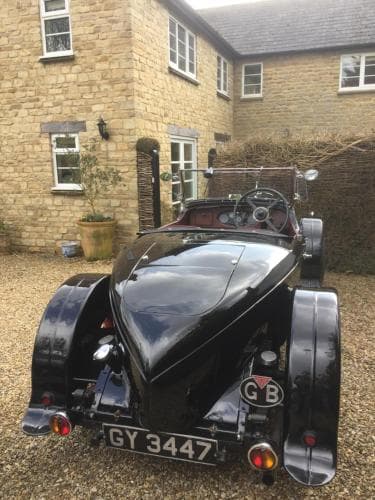 A sleek vintage black car parked on a gravel driveway in front of a charming stone house evokes a nostalgic mood, perfect for classic car enthusiasts exploring historic countryside settings.