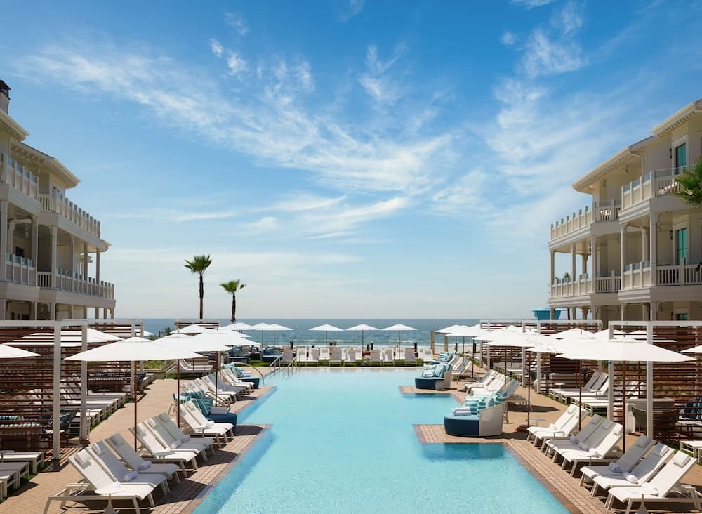 A serene coastal resort features a long pool lined with lounge chairs and umbrellas under a clear blue sky, ideal for luxury beachside relaxation.