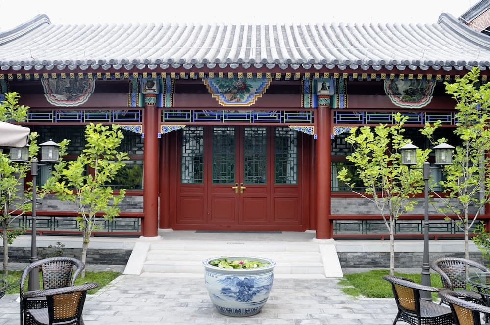 A serene courtyard features a traditional Chinese building with intricate red doors and tiled roof, surrounded by greenery, outdoor seating, and a decorative blue-and-white porcelain planter, ideal for cultural exploration and relaxation.