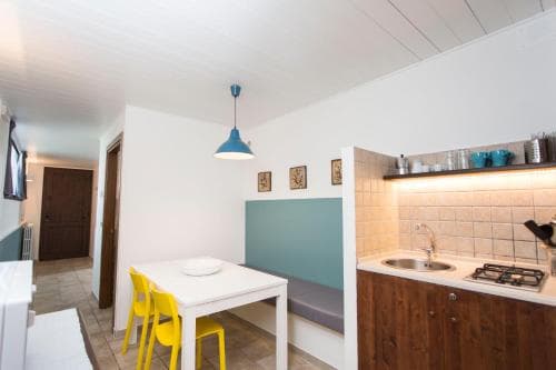 A cozy kitchenette with white walls, wooden cabinets, a small dining table with yellow chairs, and a blue pendant light creates a warm and inviting atmosphere ideal for travelers seeking a comfortable self-catering accommodation.
