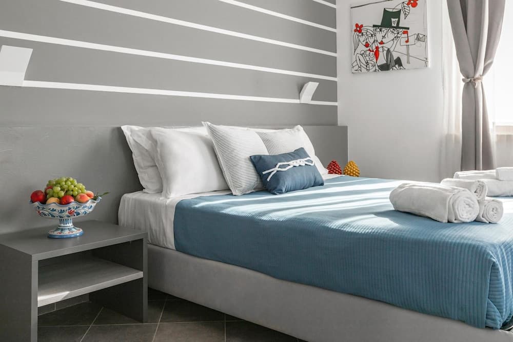 A cozy hotel room features a neatly made bed with blue linens, a fruit bowl on a modern nightstand, and soft natural light filtering through sheer curtains, ideal for travelers seeking comfort and relaxation.
