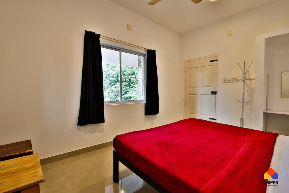 A cozy bedroom with a vibrant red bedspread, black curtains framing a window view of greenery, and minimal furnishings creates a serene atmosphere ideal for travelers seeking a peaceful retreat.