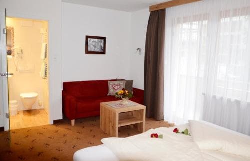 A cozy hotel room features a red sofa, wooden coffee table with flowers, sheer curtains letting in soft light, and an open bathroom door, ideal for travelers seeking comfort and convenience.